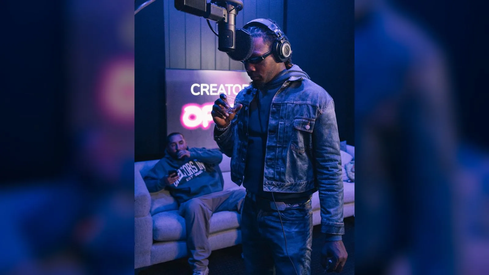 Recording artist holding a microphone in a blue-lit studio while a guest in Creators Inc merch sits on the couch