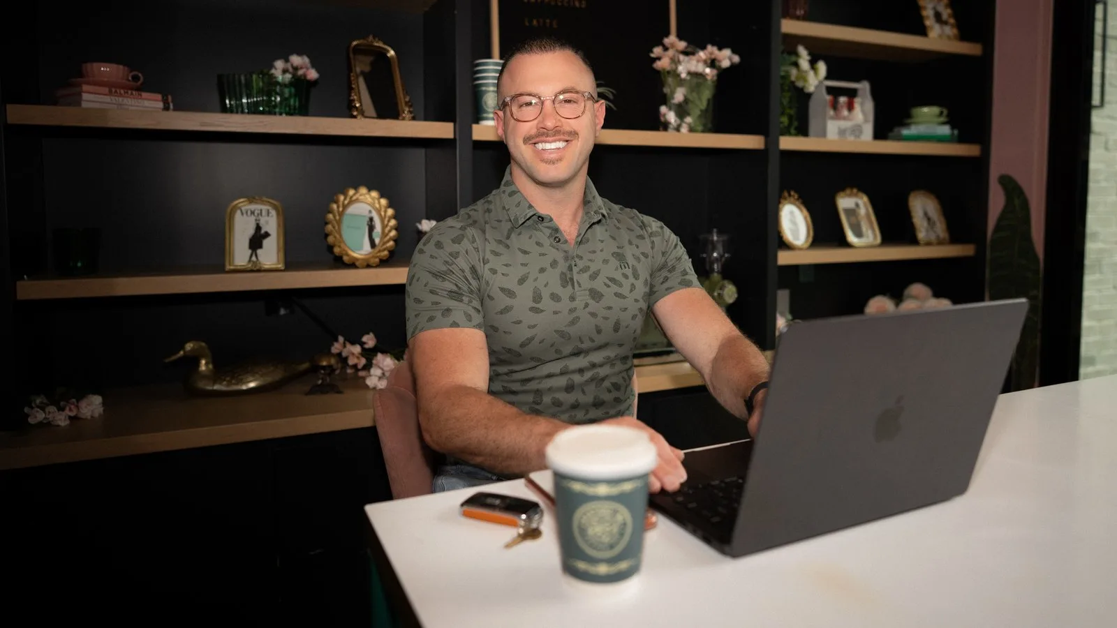 Founder Andy smiling warmly while working on a laptop at a styled coffee-bar office counter with a coffee cup beside him.