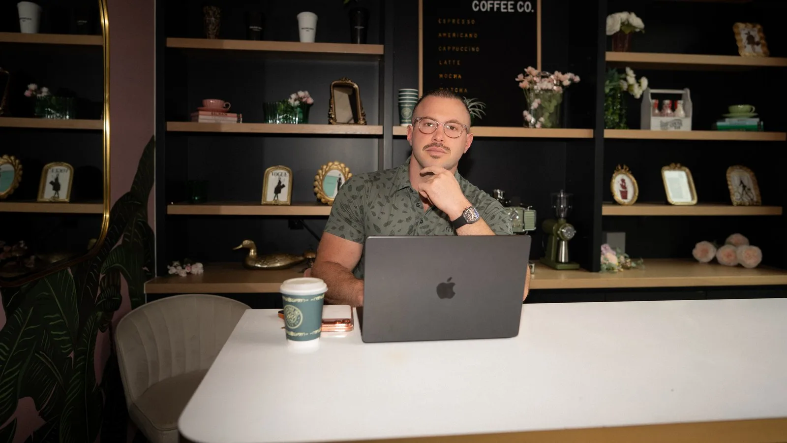 Founder Andy in a thoughtful pose with hand on chin, seated at a laptop in a styled coffee-bar office under a Coffee Co. sign.